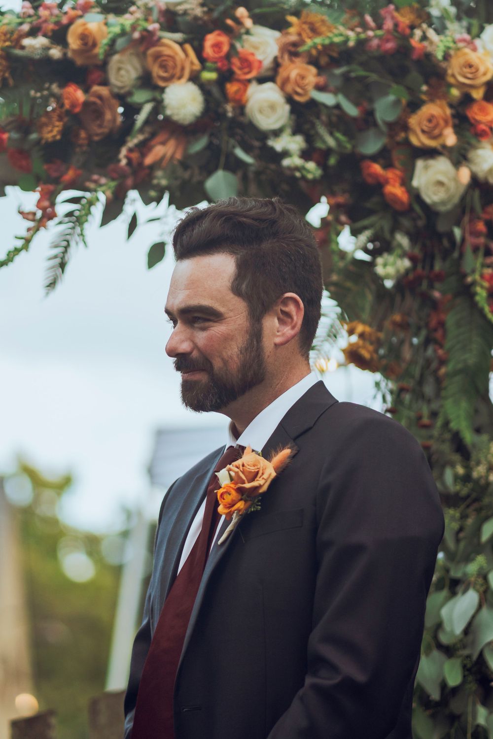 A groom hears vows from his soon to be wife in a black and red tuxedo with a flowery backdrop, capturing a romantic wedding scene