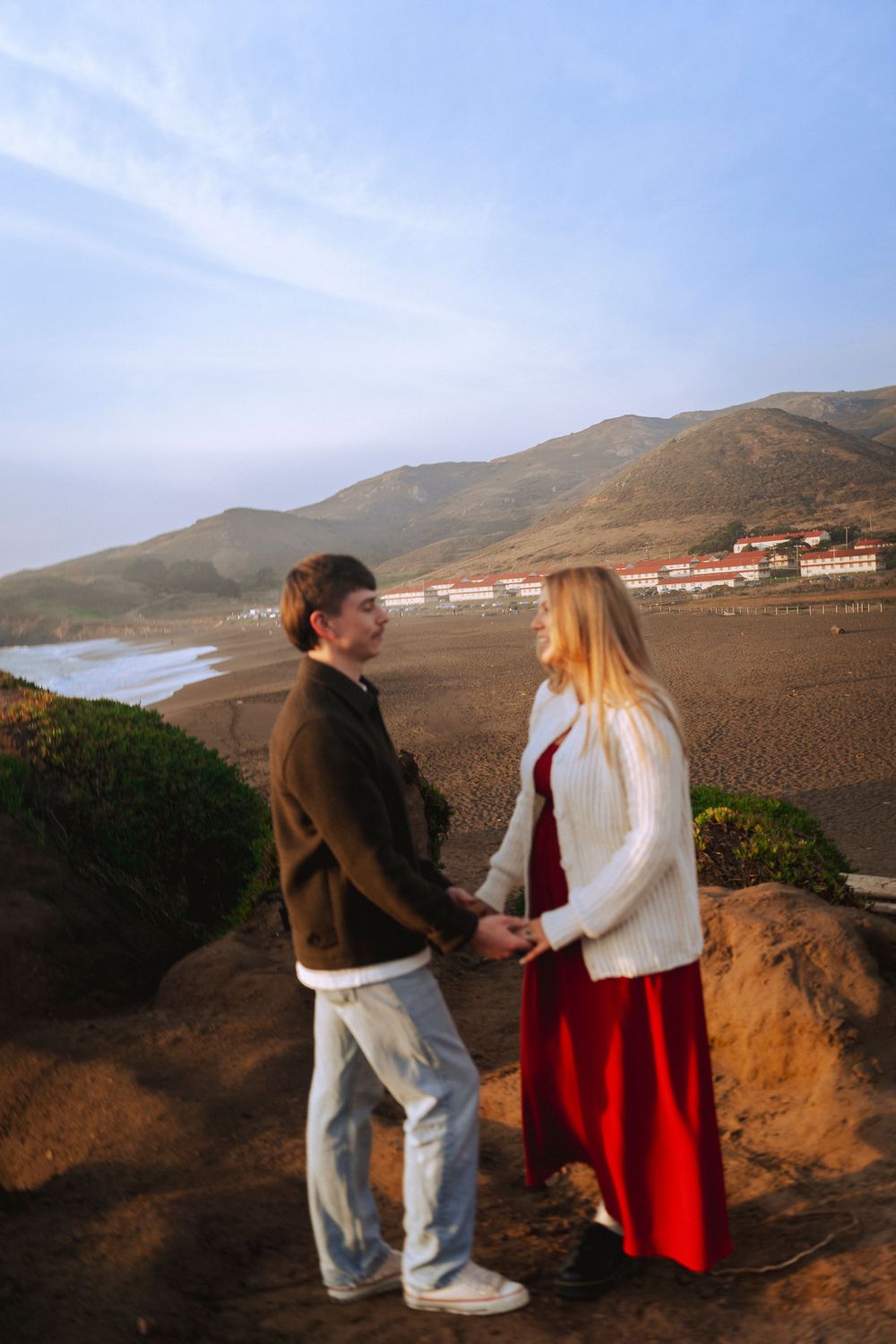 A couple stands on a sandy beach at sunset, holding hands, with green hills behind them. A warm, romantic moment captured in golden light.
