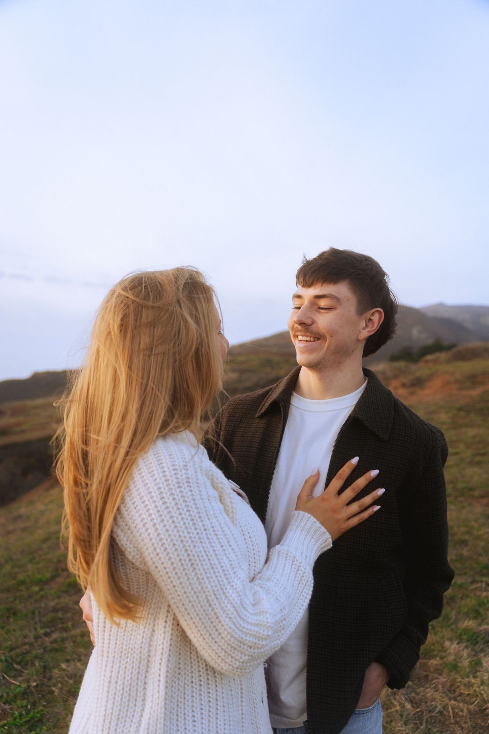 A couple stands on a sandy beach at sunset, holding hands, with green hills behind them. A warm, romantic moment captured in golden light.
