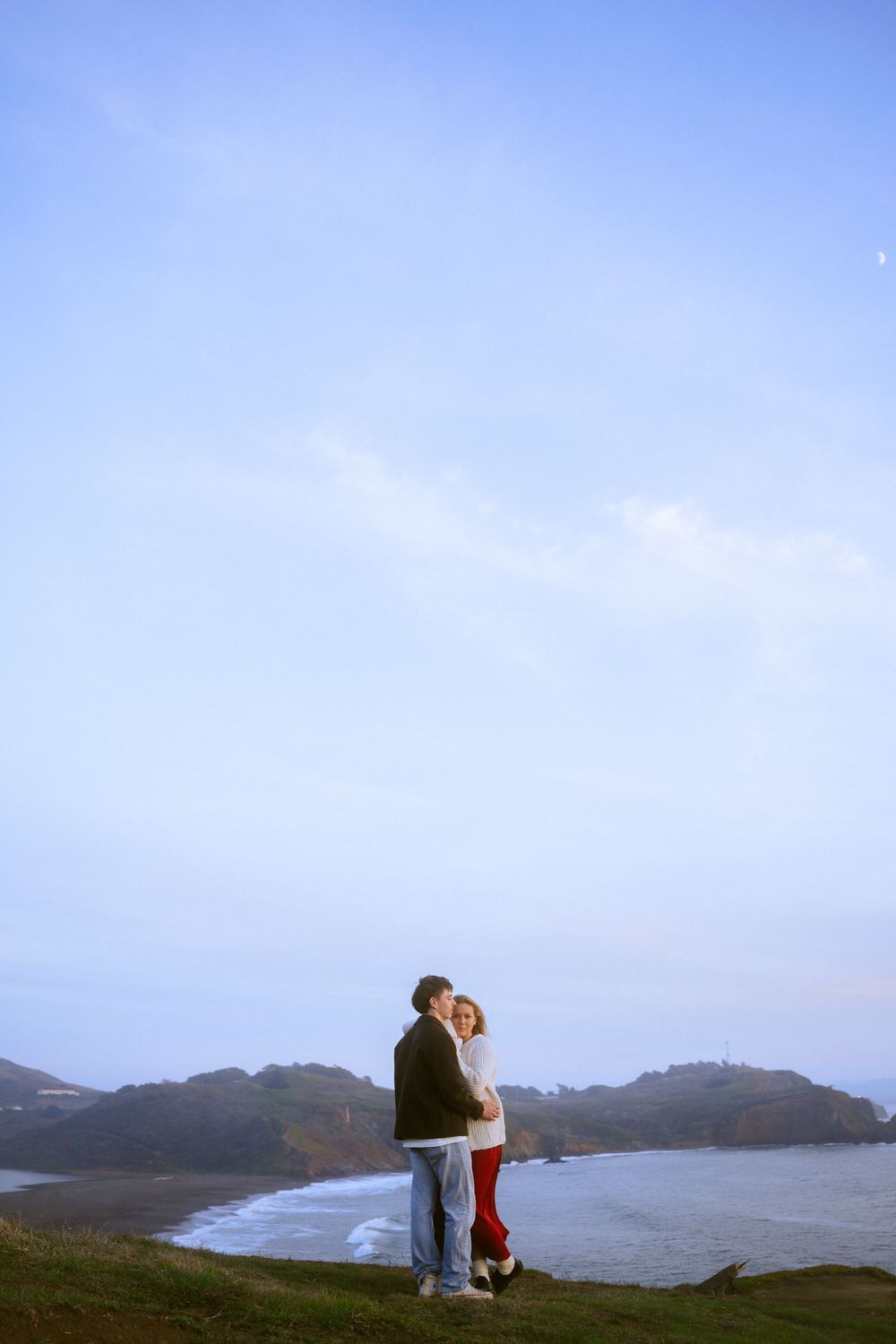 A couple stands on a sandy beach at sunset, holding hands, with green hills behind them. A warm, romantic moment captured in golden light.