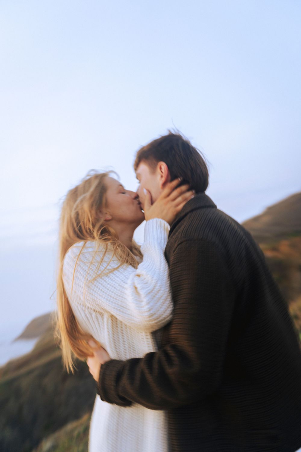 A couple stands on a sandy beach at sunset, holding hands, with green hills behind them. A warm, romantic moment captured in golden light.