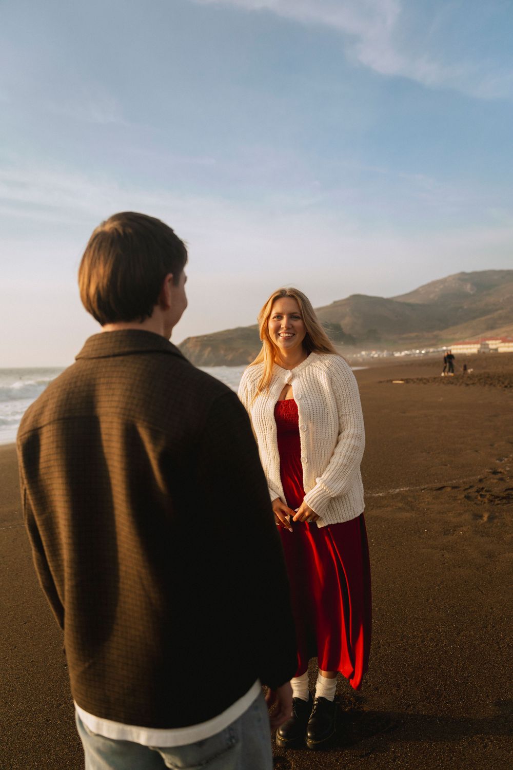 A couple stands on a sandy beach at sunset, holding hands, with green hills behind them. A warm, romantic moment captured in golden light.