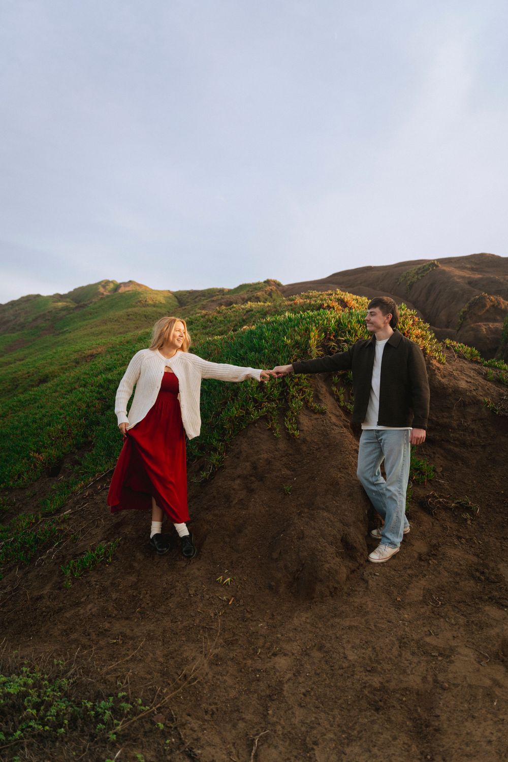 A couple stands on a sandy beach at sunset, holding hands, with green hills behind them. A warm, romantic moment captured in golden light.