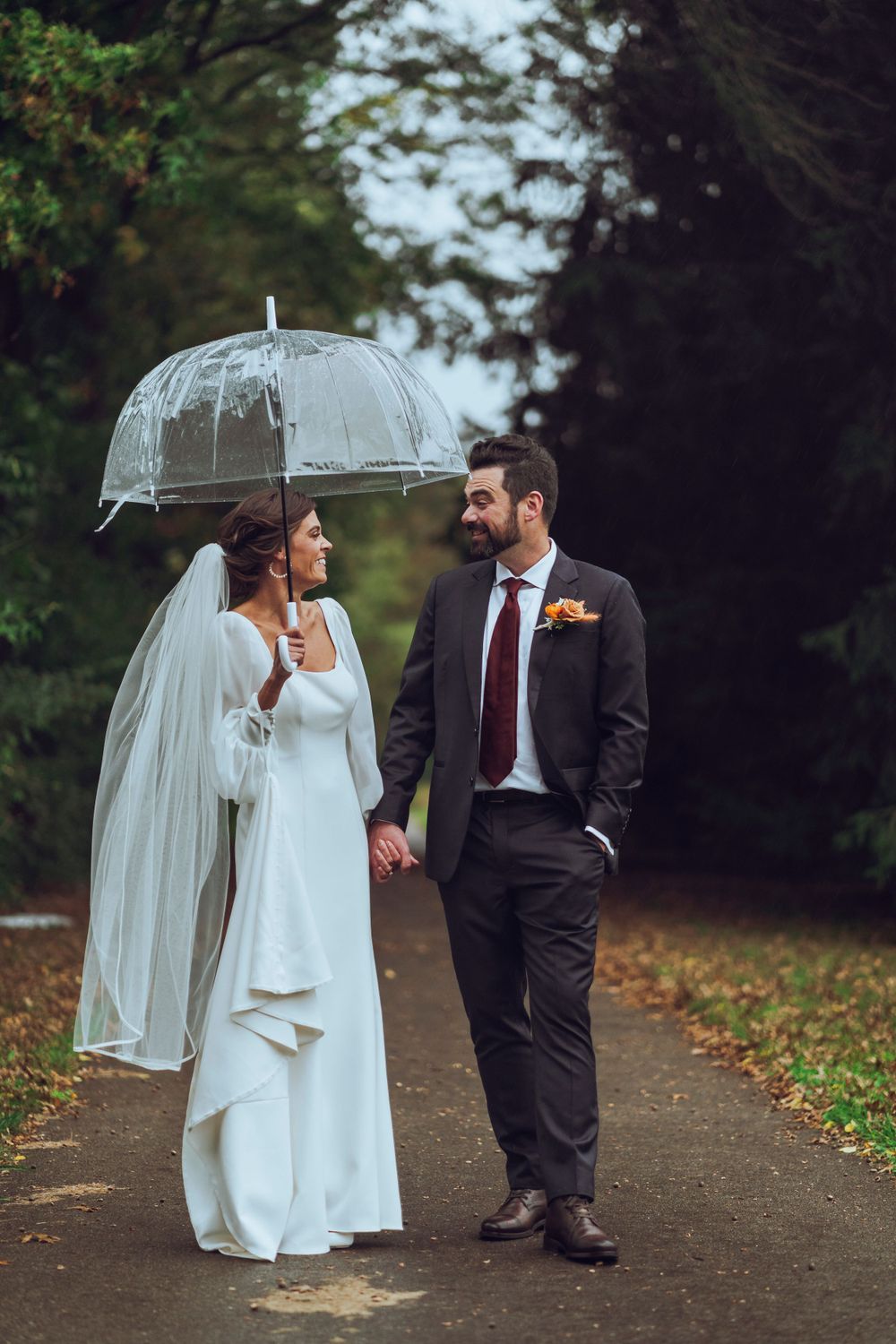 A bride and groom walk hand in hand in the rain, smiling under clear umbrellas, capturing a romantic and joyful wedding moment.