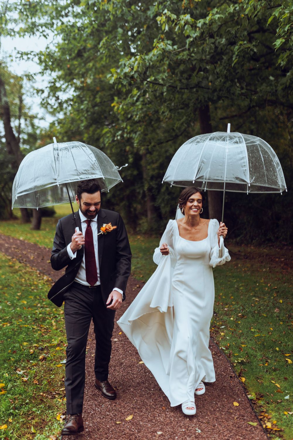 A bride and groom walk hand in hand in the rain, smiling under clear umbrellas, capturing a romantic and joyful wedding moment.