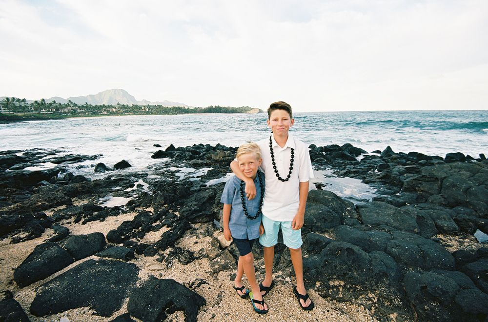 Two people standing on black volcanic rocks with ocean waves behind them.
