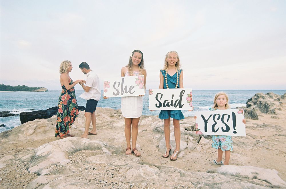 Beach proposal scene with people holding signs on rocky coastline.