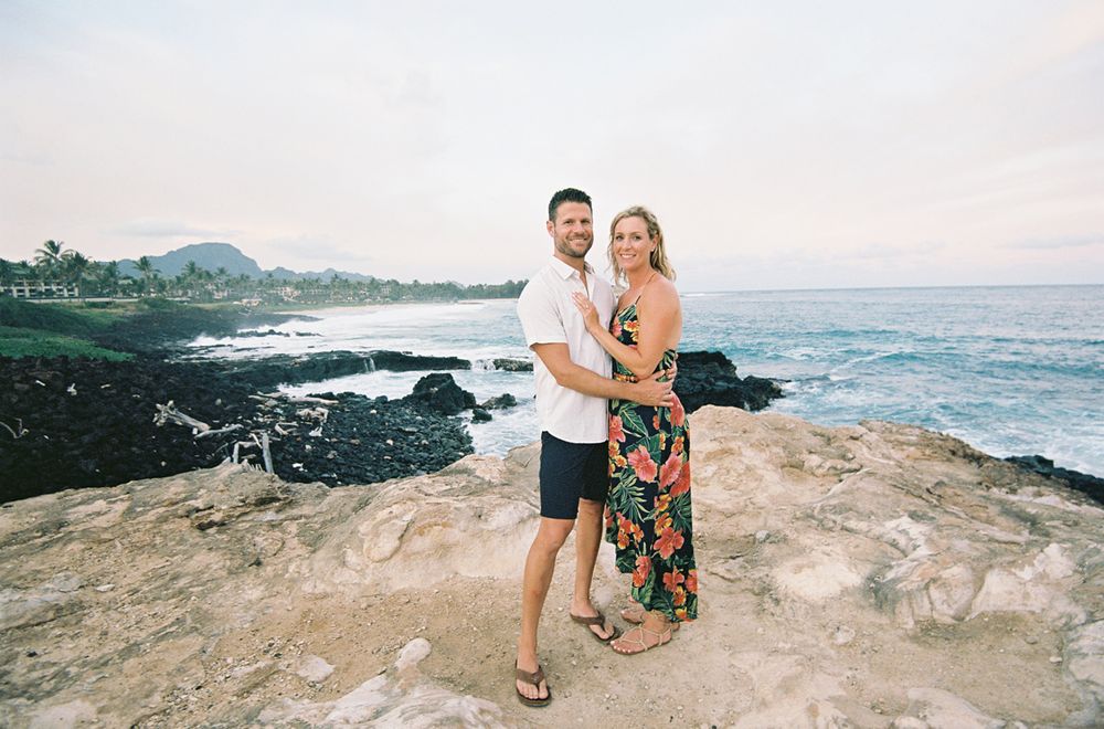 Couple posing together on volcanic rocks with ocean waves and coastline in background.