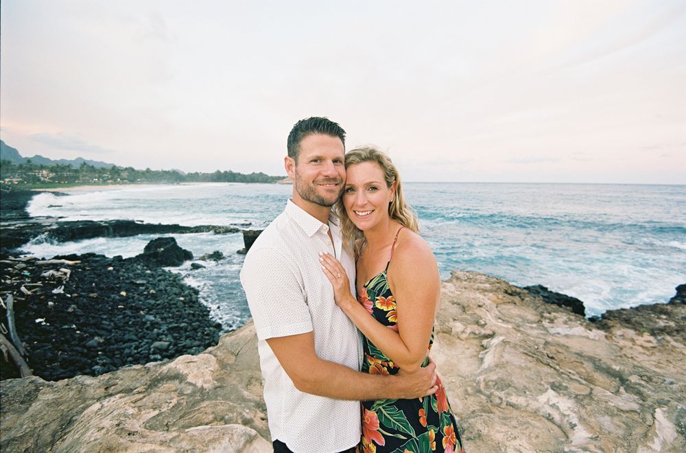 Romantic couple embracing on rocky coastal shore at sunset with ocean waves crashing in the background.