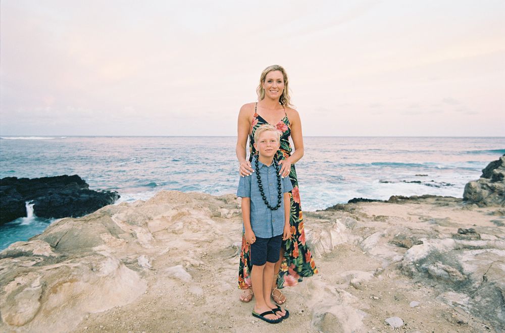 Woman in floral dress standing on rocky ocean overlook during sunset.