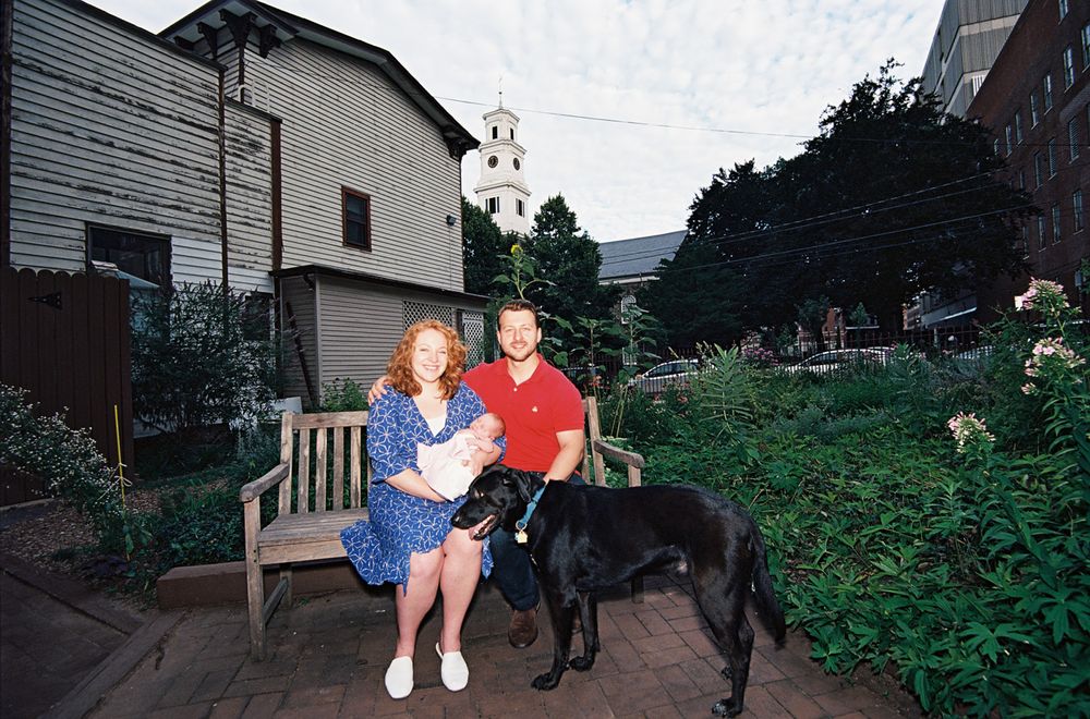 Couple sitting on garden bench with black dog surrounded by lush greenery.