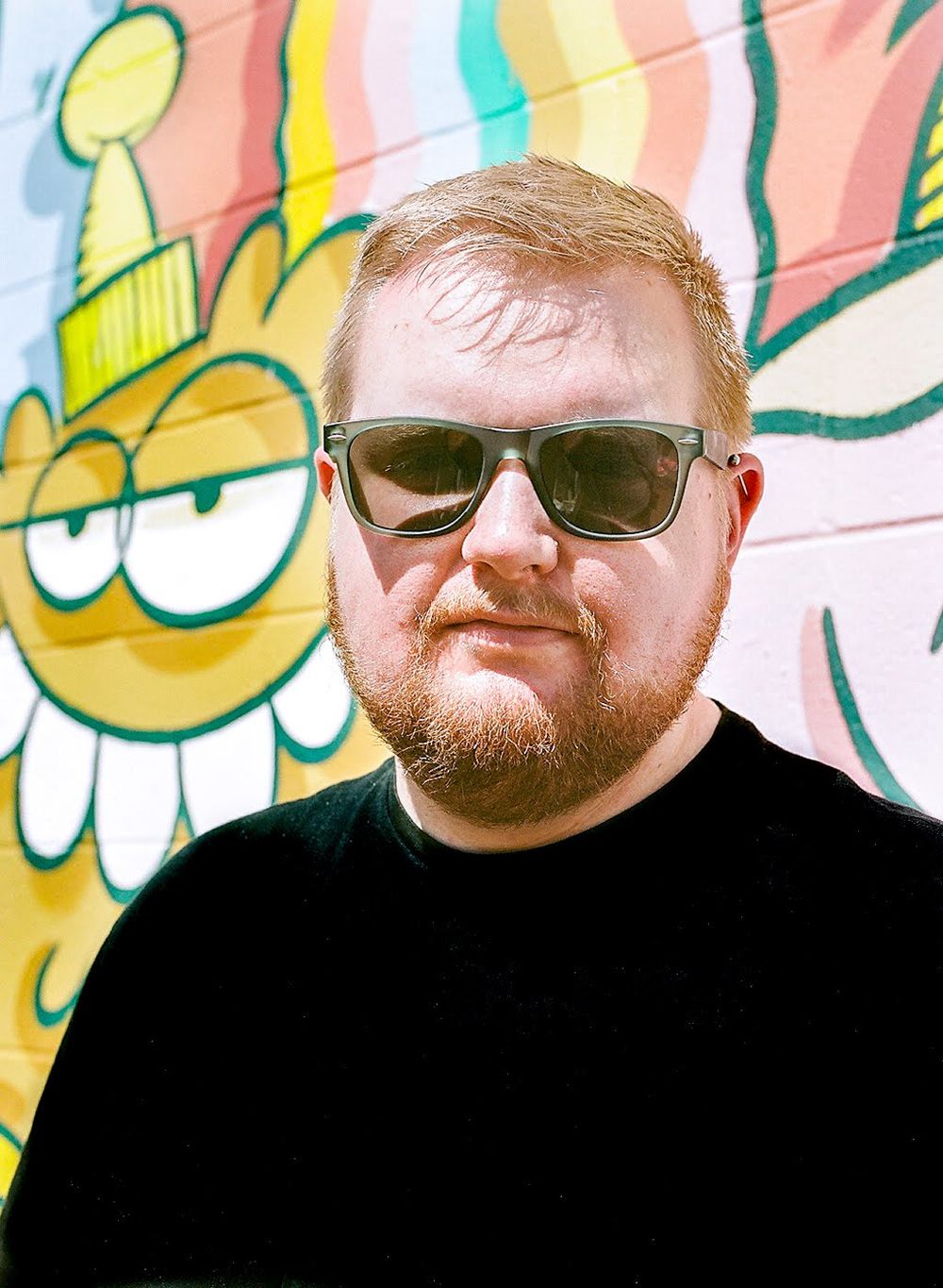 A person wearing sunglasses and black shirt poses in front of vibrant street art mural.