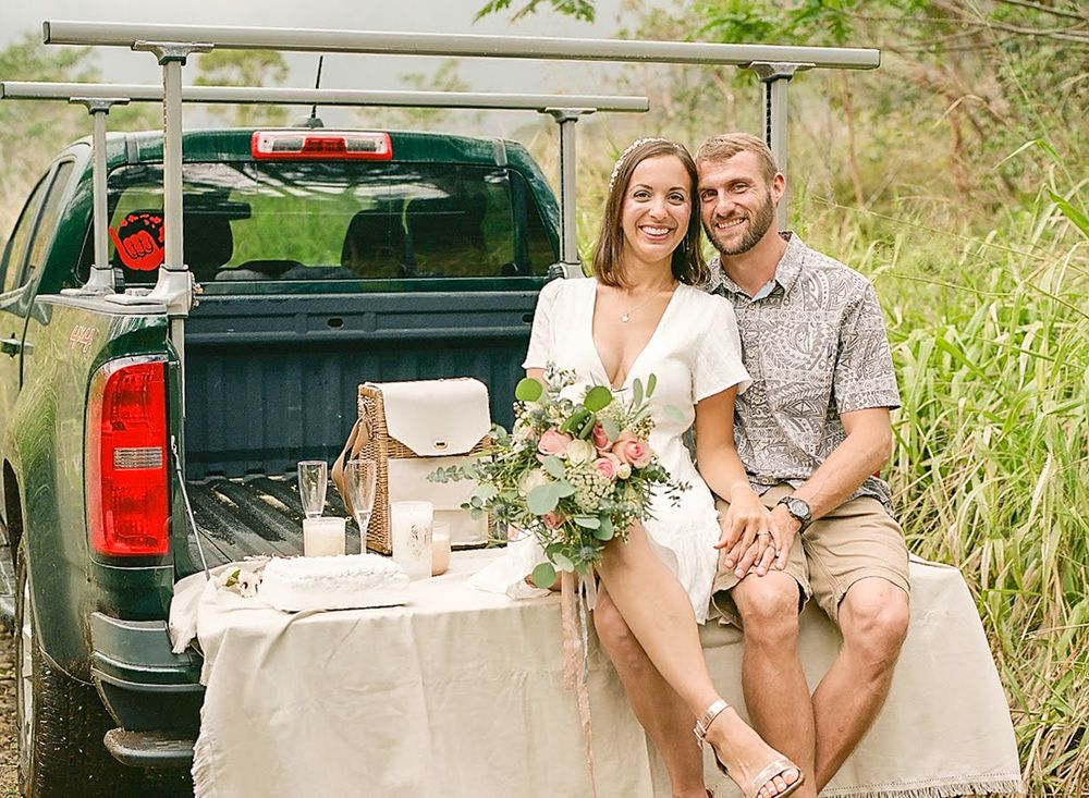 A couple sits on the tailgate of a pickup truck with flowers and outdoor scenery.