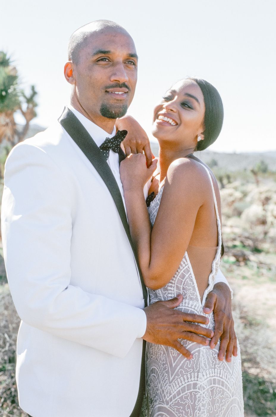 A romantic bride and groom embrace outdoors in formal wedding attire against a desert backdrop.