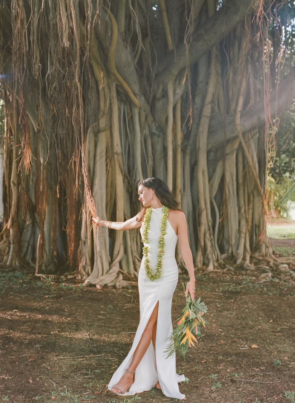 Bride in white dress with lei stands against dramatic banyan tree backdrop in tropical setting.