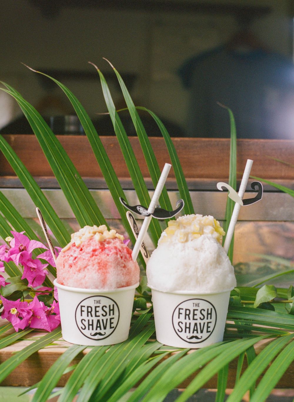 Two tropical shaved ice desserts with pink and white toppings decorated with palm leaves.