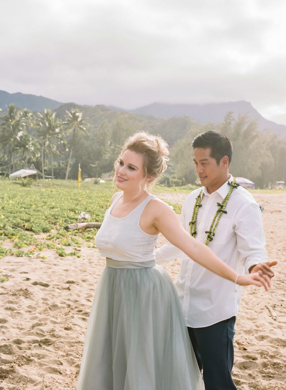 Wedding couple dances on beach with mountains in background during overcast day.