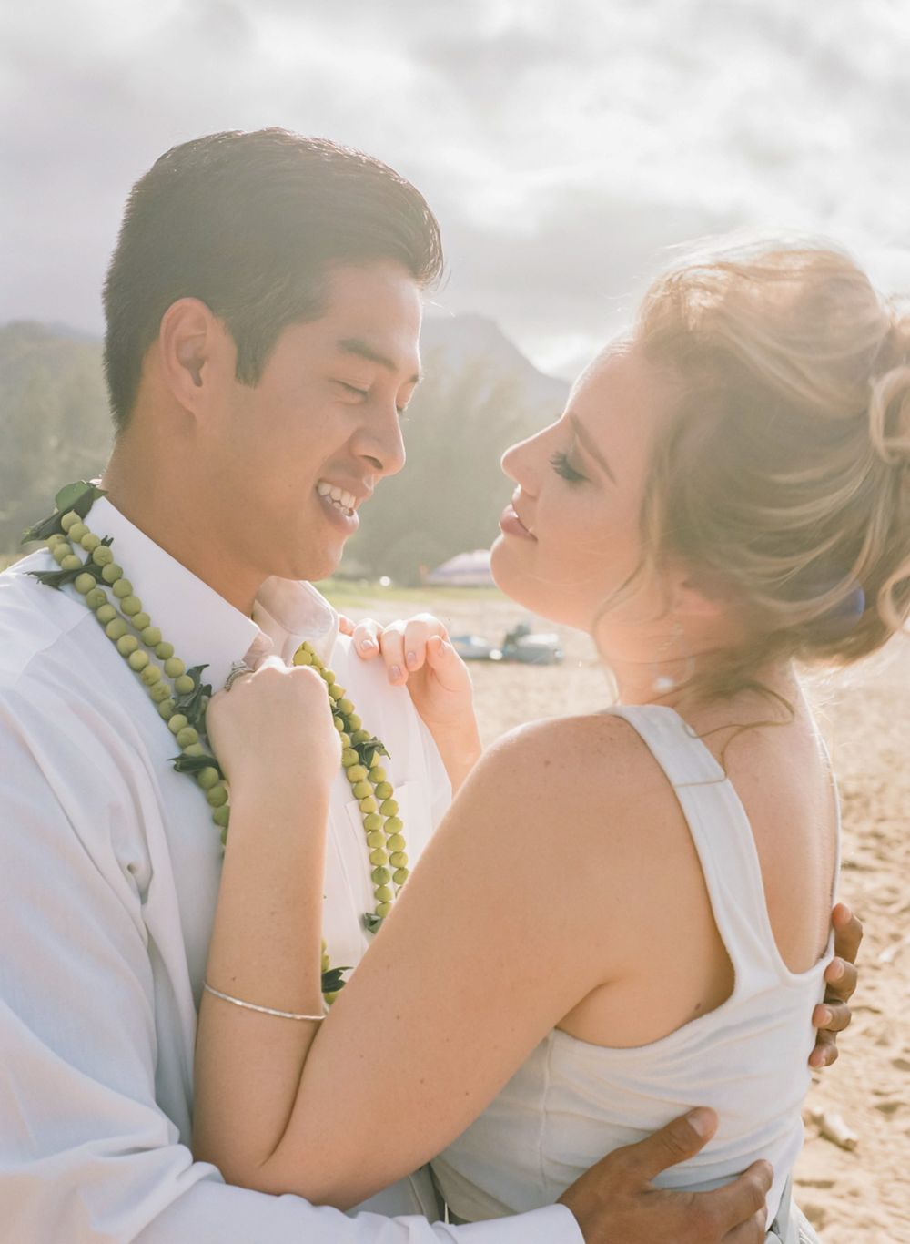 Couple shares intimate moment during beach wedding photoshoot with mountains in background.