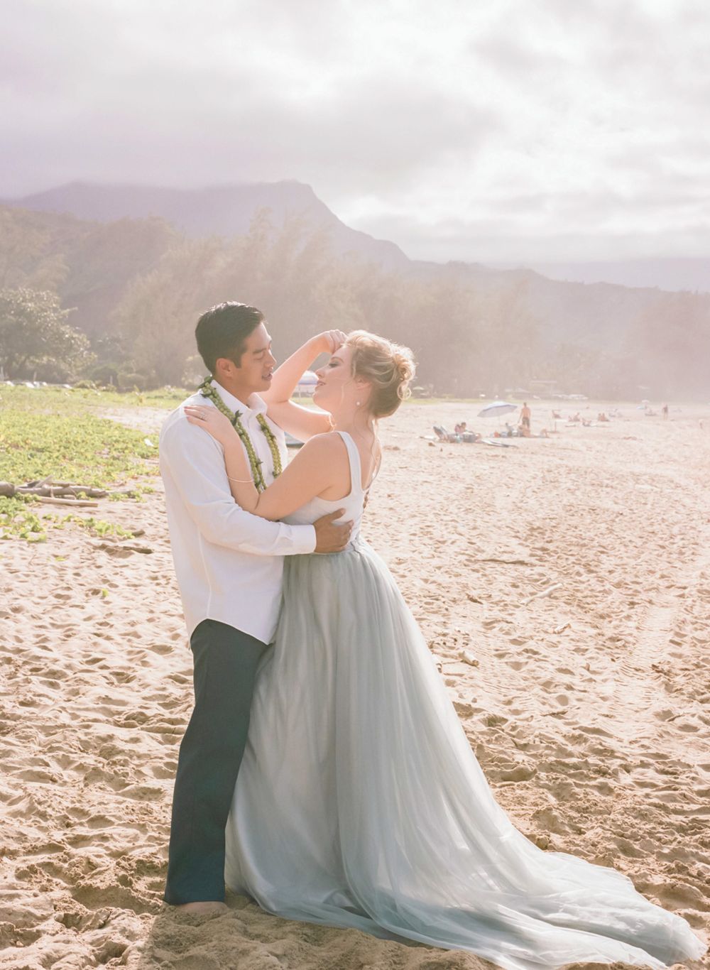 Romantic couple embraces on a Hawaiian beach with mountains in background during sunset wedding photoshoot.