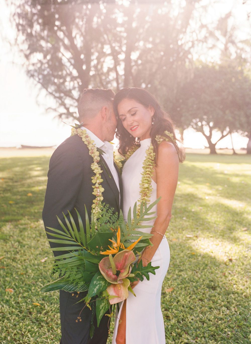 Newlyweds wearing traditional Hawaiian leis embrace in sunlit garden.