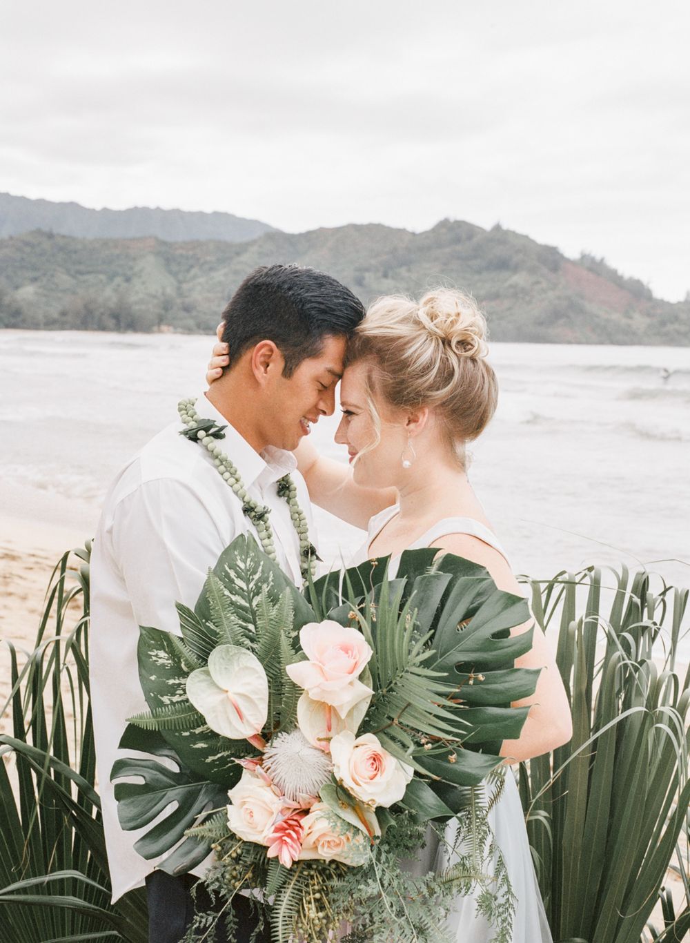 Intimate moment between couple with tropical bouquet against mountainous beach backdrop.