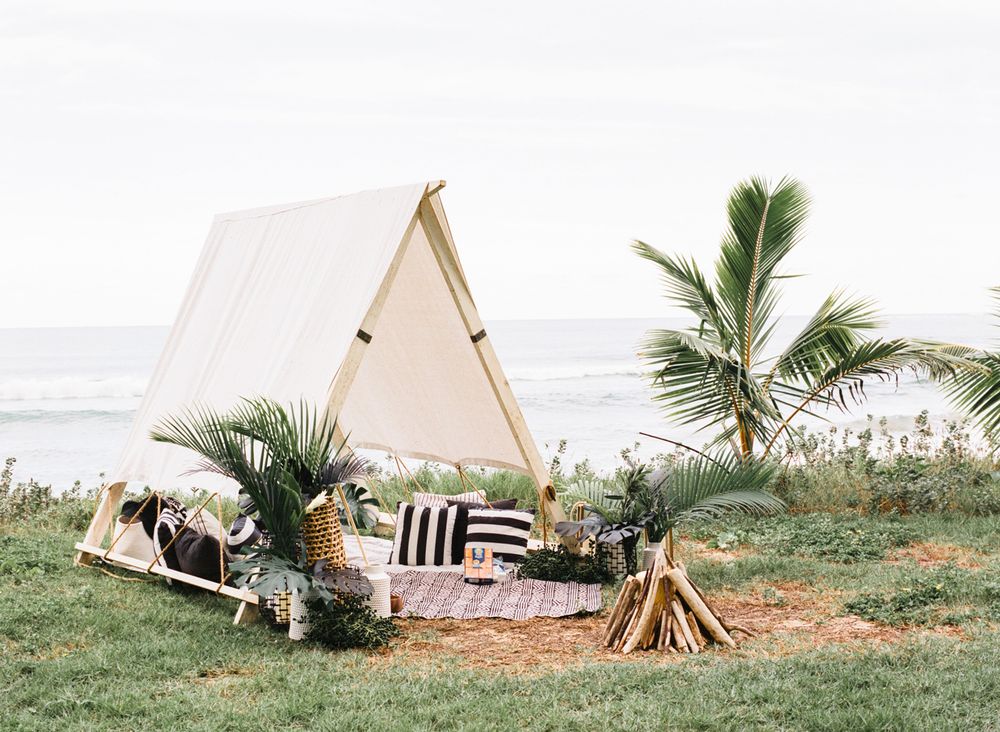 A-frame tent setup on grass with palm trees and beach views featuring pillows and blankets for glamping.