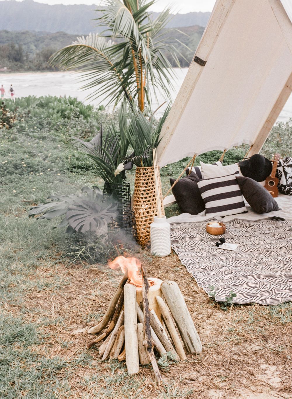 Boho beach picnic setup with A-frame tent, striped pillows and driftwood campfire.