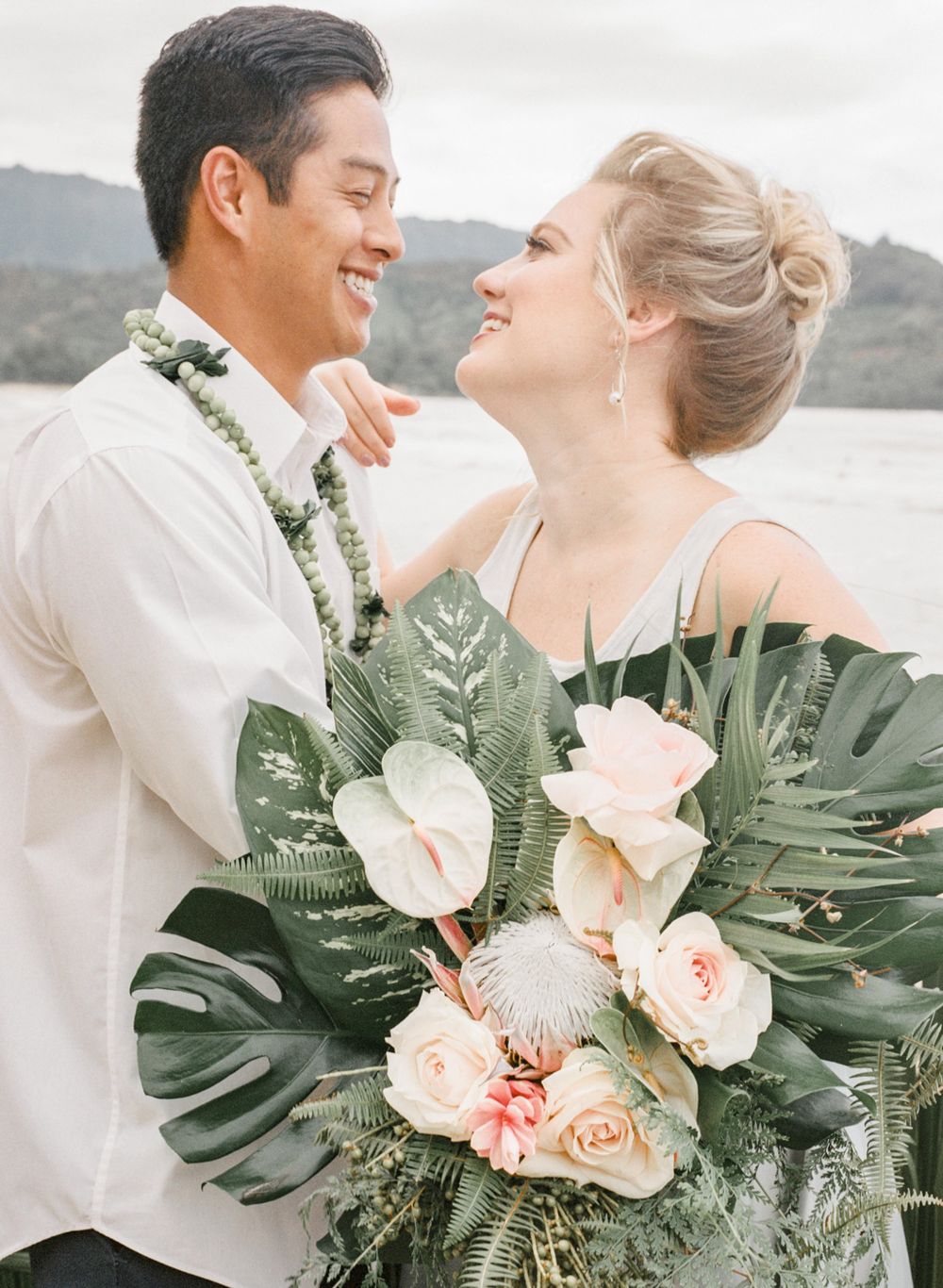 Tropical wedding portrait with monstera leaves and peach roses against ocean backdrop.