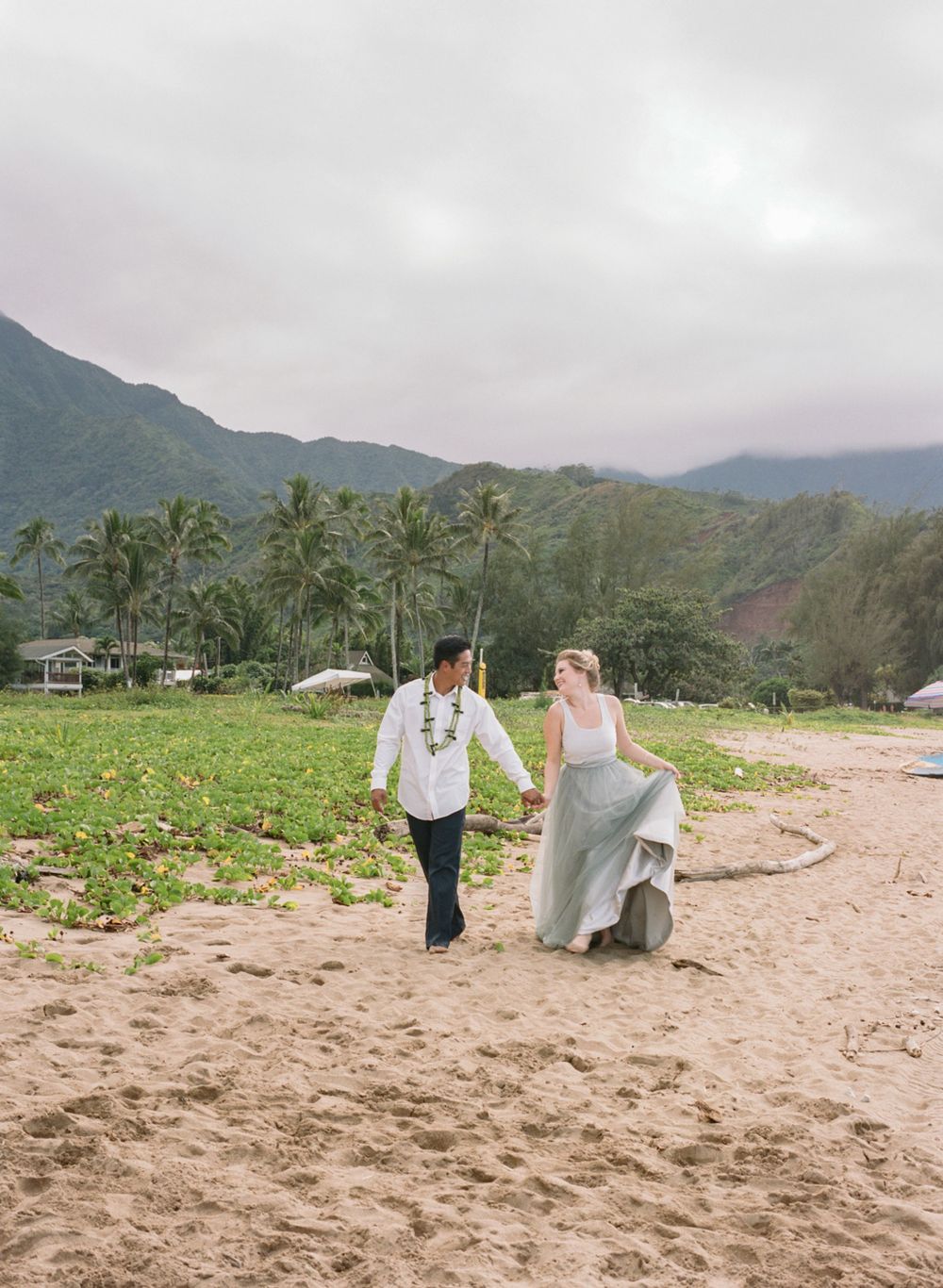 Couple walking along Hawaiian beach with mountains and palm trees in background.