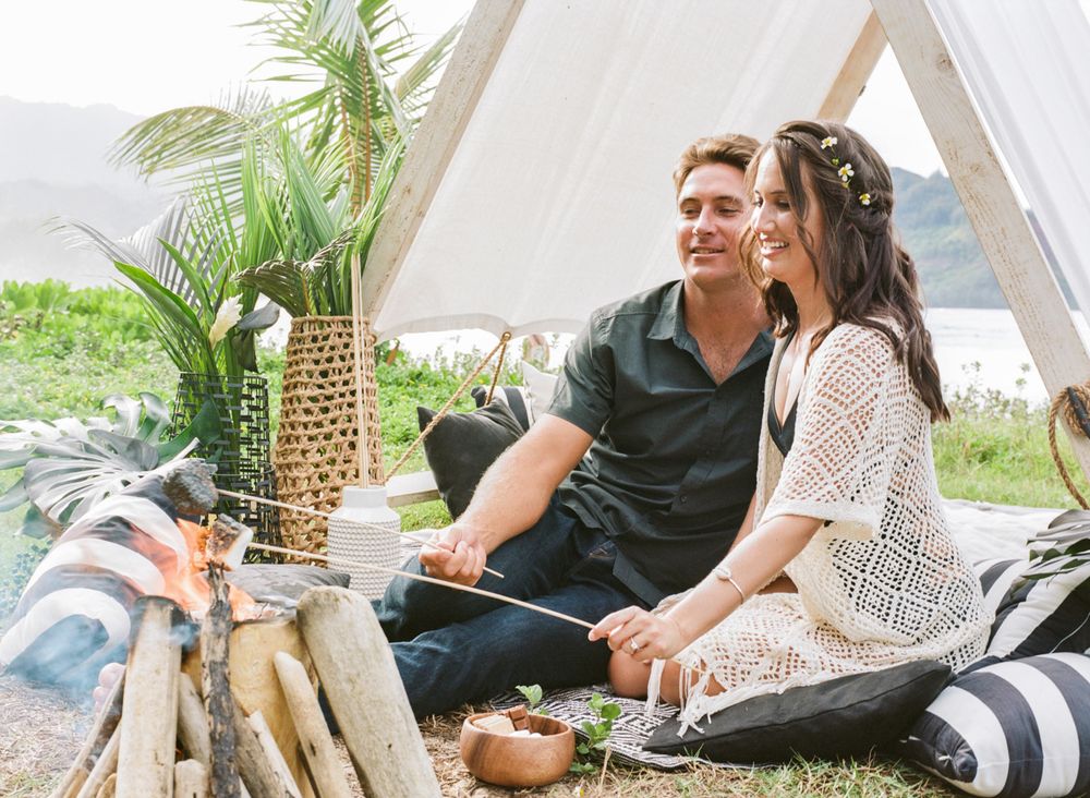 Cozy glamping setup with couple lounging by A-frame tent surrounded by tropical decor.