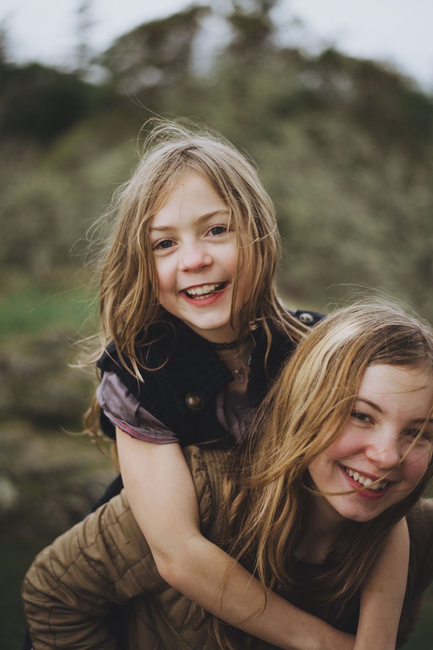 Two smiling sisters enjoy a playful piggyback ride outdoors, with wind-swept hair and joyful expressions.