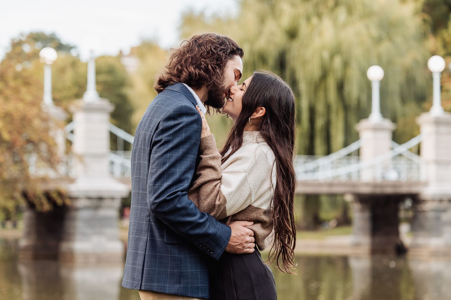A romantic kiss by a lake with a white bridge and willow trees in the background.