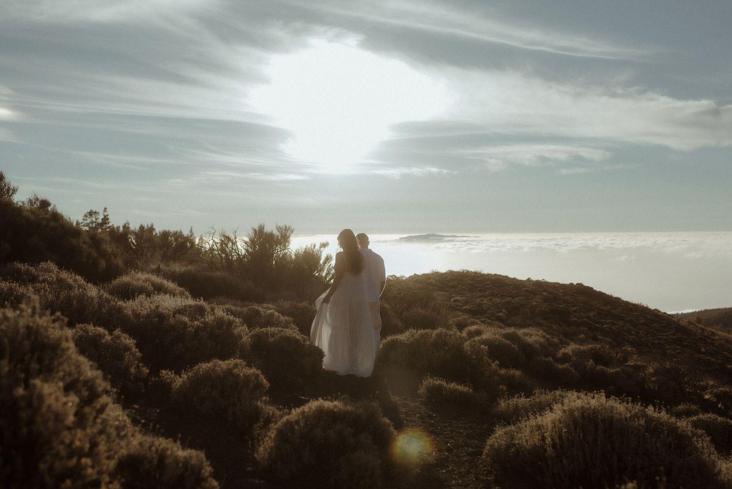 A wedding couple stands silhouetted against the setting sun on a mountain trail. Tenerife Wedding Photographer at Teide.