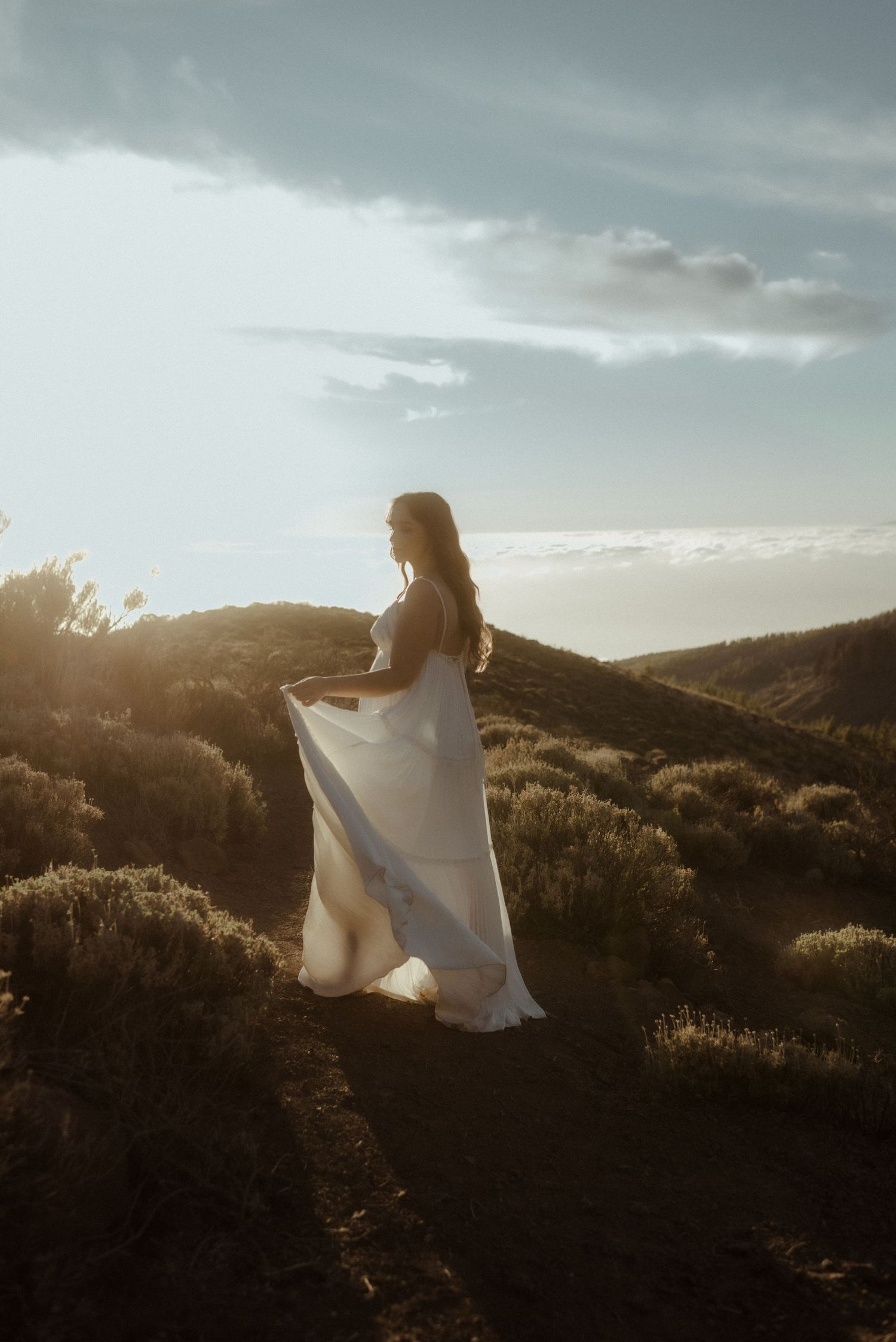 A bride in a flowing white dress poses on a hilltop during sunset. Tenerife Wedding Photographer at Teide.
