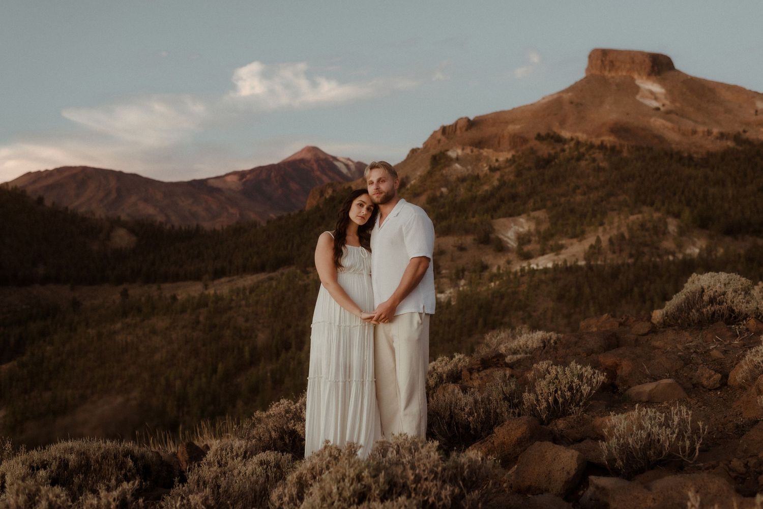 Couple embraces on mountain ridge at sunset wearing ethereal white outfits against rugged landscape. Tenerife Wedding Photographer at Teide.