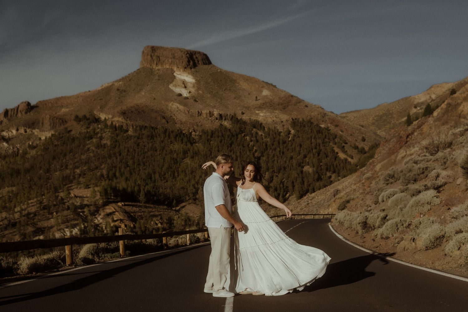 A couple in wedding attire stands on a mountain road with dramatic cliffs and peaks in the background. Tenerife Wedding Photographer