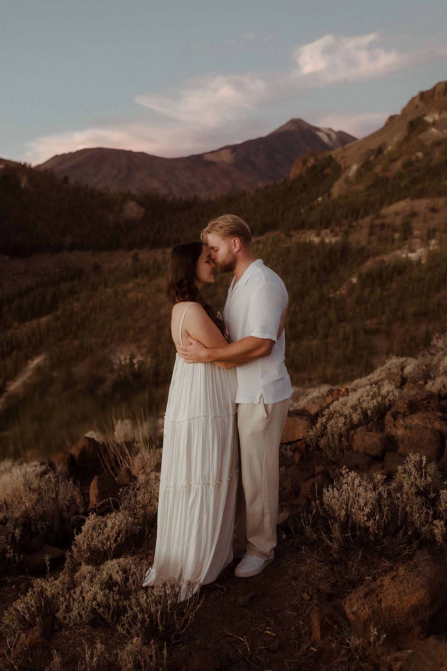 Soft sunset lighting illuminates couple in flowing white attire on mountain trail. Tenerife Wedding Photographer at Teide.