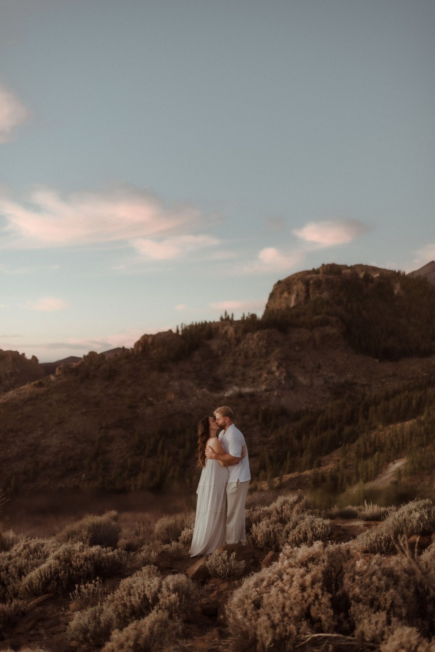 Couple stands together on hillside with dramatic cliffs and evening sky behind them. Tenerife Wedding Photographer at Teide.