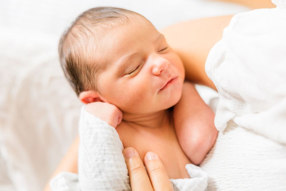 A peaceful sleeping newborn baby wrapped in soft white blankets rests gently against a light background.