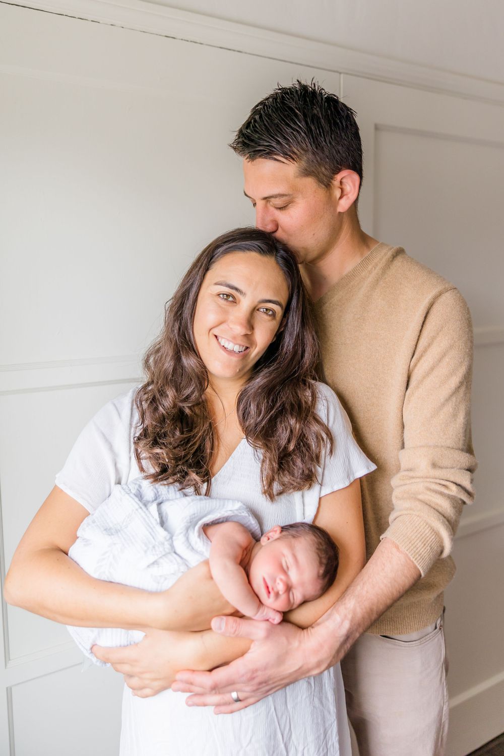 A couple in casual attire sharing an intimate moment while holding their sleeping newborn against a white wall.