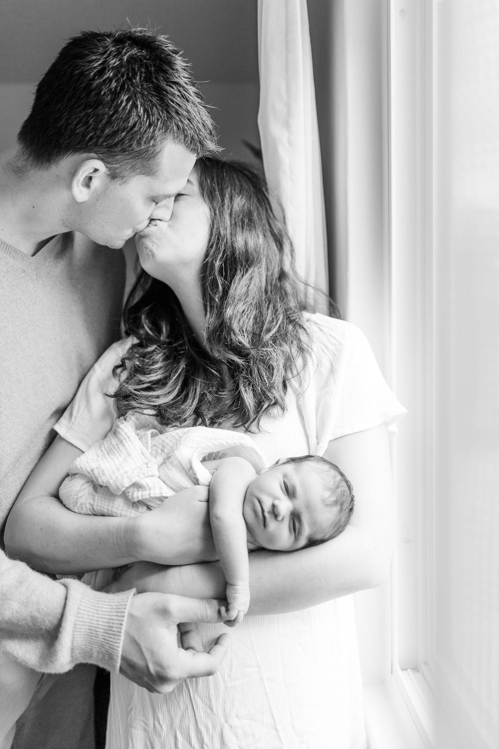 A tender black and white portrait of new parents sharing an intimate moment with their sleeping newborn by a window.