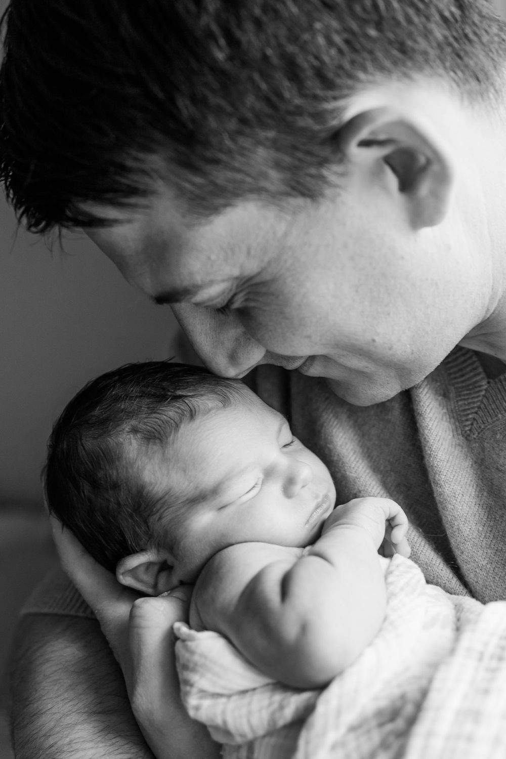 An intimate black and white portrait of a sleeping newborn being cradled close while receiving a gentle kiss.