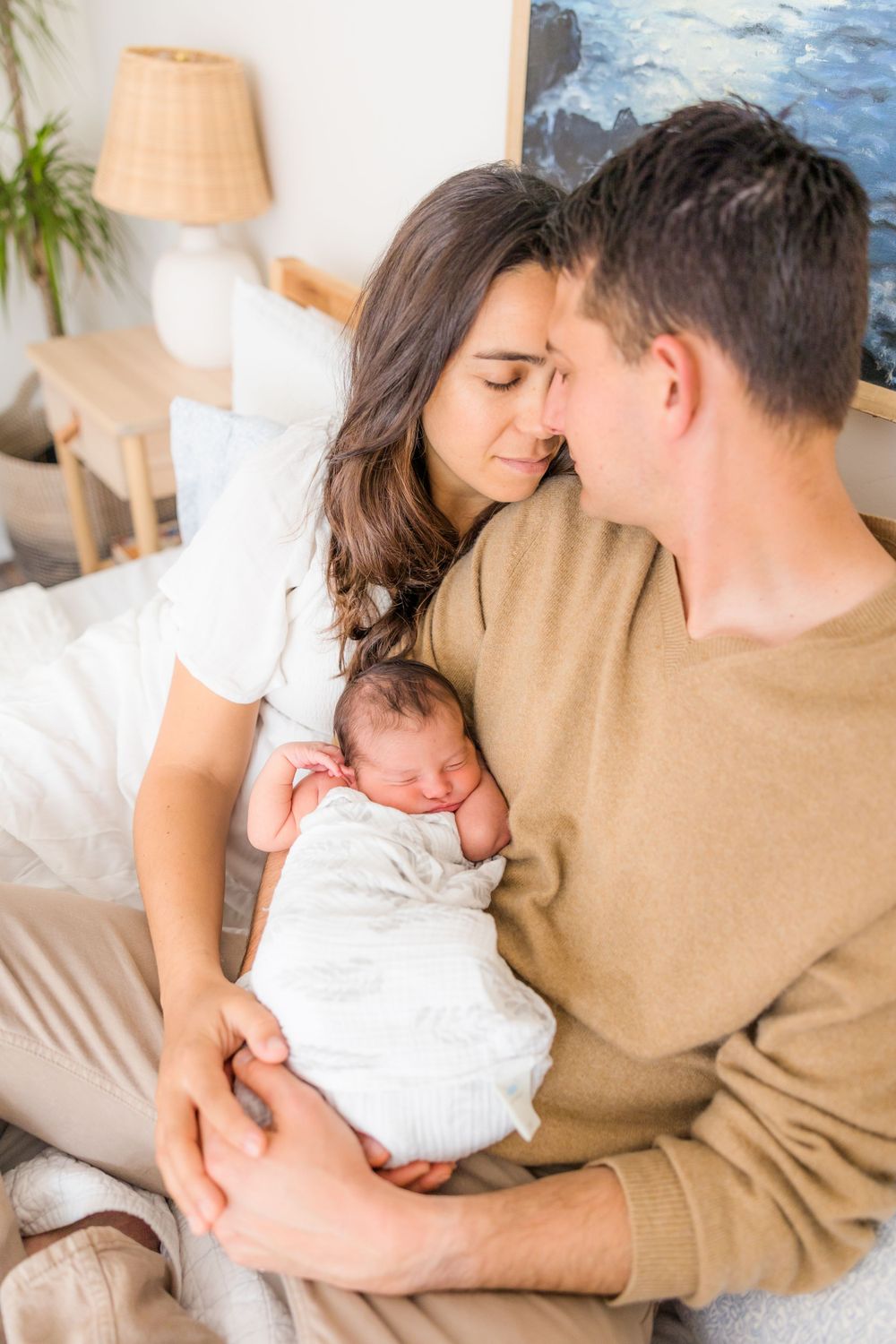 A tender family moment as new parents gaze lovingly at their sleeping newborn baby wrapped in white on a couch.
