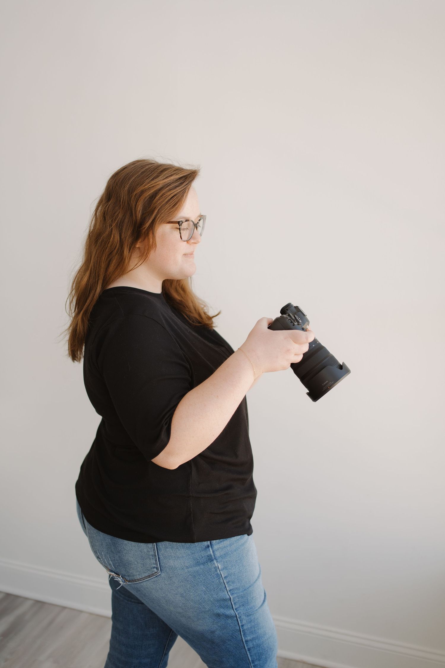A photographer in a black shirt and jeans checks their camera while standing against a white wall in a minimal setting.