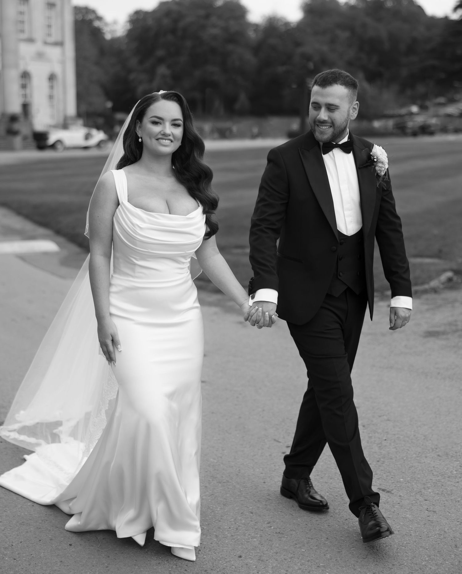 A couple walks hand in hand on their wedding day, with the bride in white gown and groom in black tuxedo.