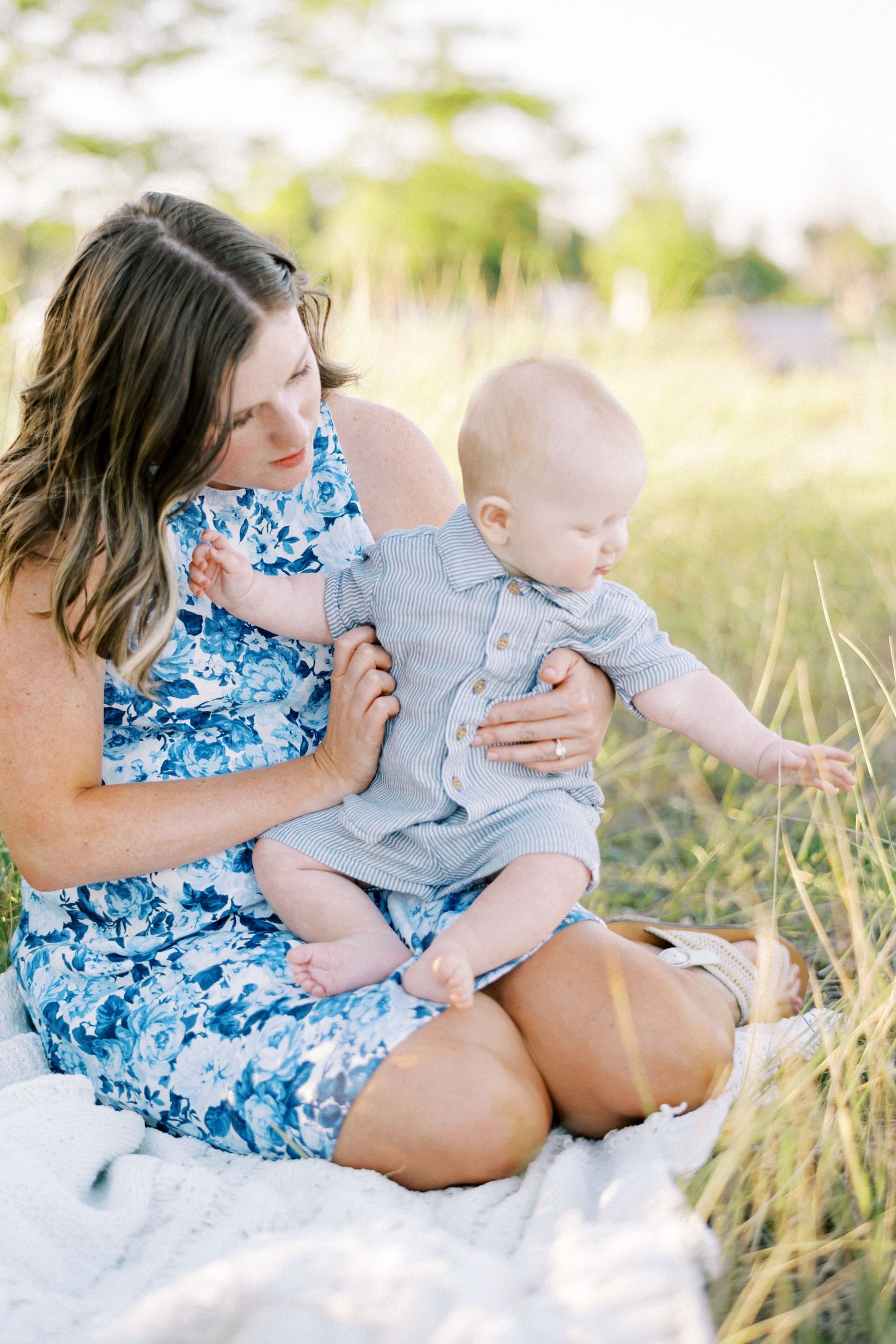 A woman in a blue floral dress sits on a blanket with a baby in gray clothing during a sunny outdoor photo session.