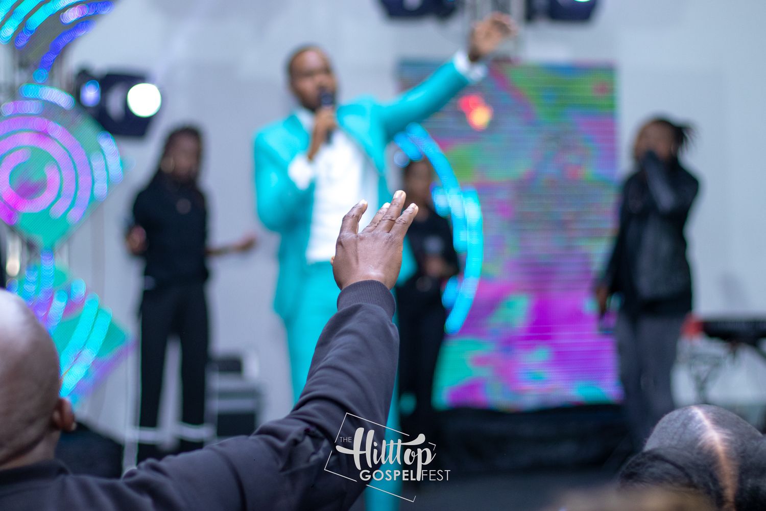 A raised hand in the foreground reaches toward a performer in a bright turquoise suit on stage with colorful lighting effects.