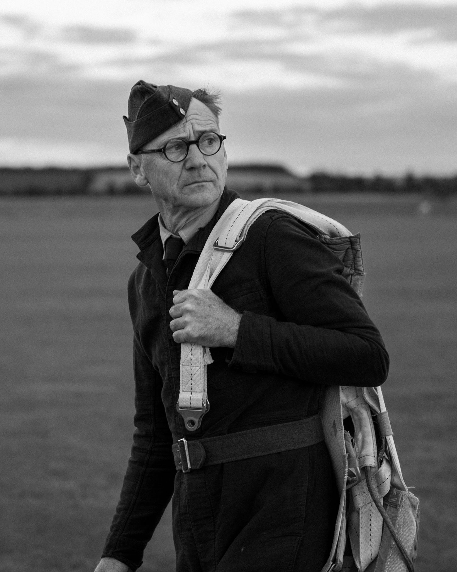 A figure in vintage aviator gear with a parachute pack stands on an airfield.