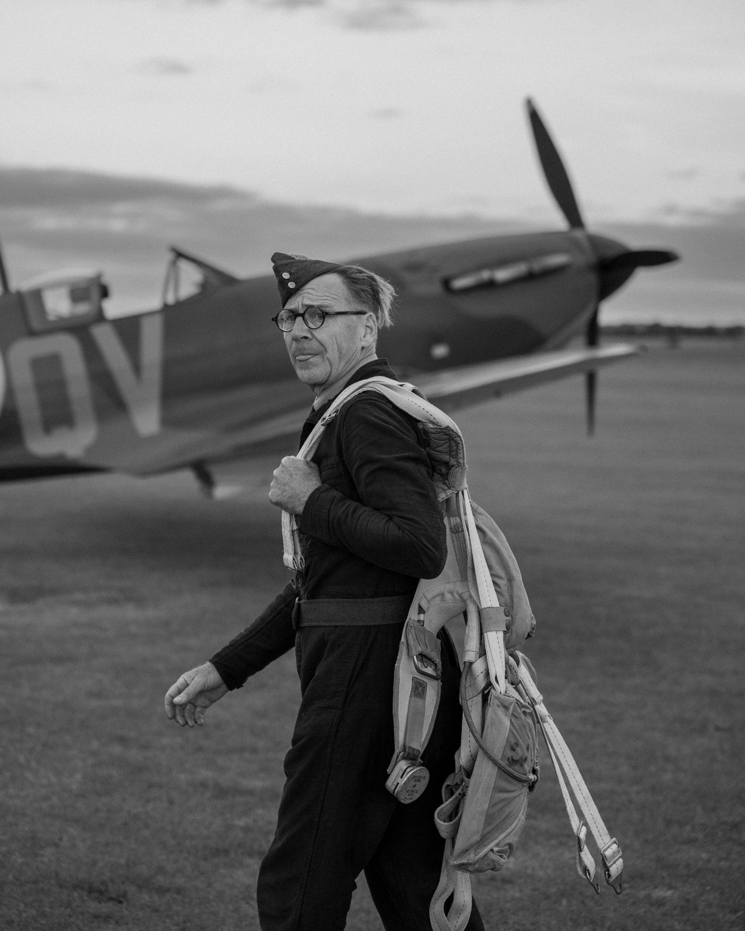 A person in flight gear walks past a parked Spitfire aircraft on an airfield.