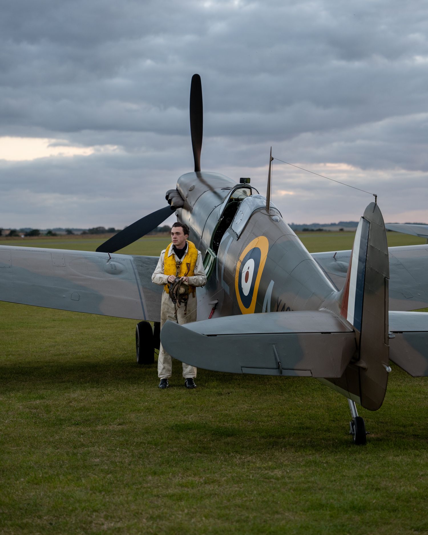 Ground crew member in yellow vest inspects a vintage Spitfire aircraft at dusk.
